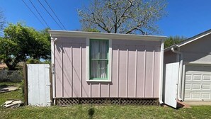 A single-story, light-colored shed with a green-framed window, a small porch area, and a garage door in the background.