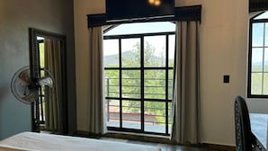 Una habitación de hotel con una cama, un ventilador de pie, un ventanal con cortinas y un televisor montado en la pared.