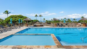 Ein Pool mit Liegestühlen und Sonnenschirmen, umgeben von Palmen und blauem Himmel.