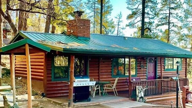 A rustic cabin with a green metal roof, wooden walls, and a chimney. There is a grill outside and a porch with chairs. - Pinetop Vista - The Wildlife Cabin (Pinetop)