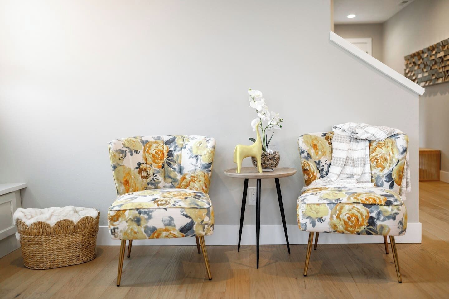 Dos sillones con tapicería floral, una mesita redonda con un jarrón de flores y una cesta tejida en el suelo.