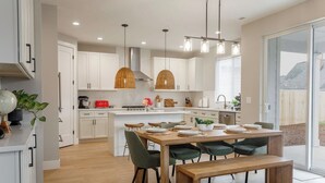 A modern kitchen with white cabinetry, a central island, and a dining area with a wooden table and chairs. - Evergreen Dream-massage chair, EV, 5tvs,3 king bed (Vancouver)