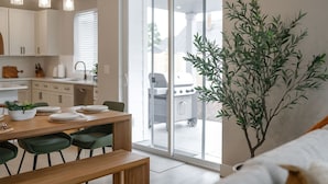 A modern kitchen with white cabinets, a dining table with green chairs, and a large sliding glass door leading to an outdoor area with a barbecue grill. - Evergreen Dream-massage chair, EV, 5tvs,3 king bed (Vancouver)
