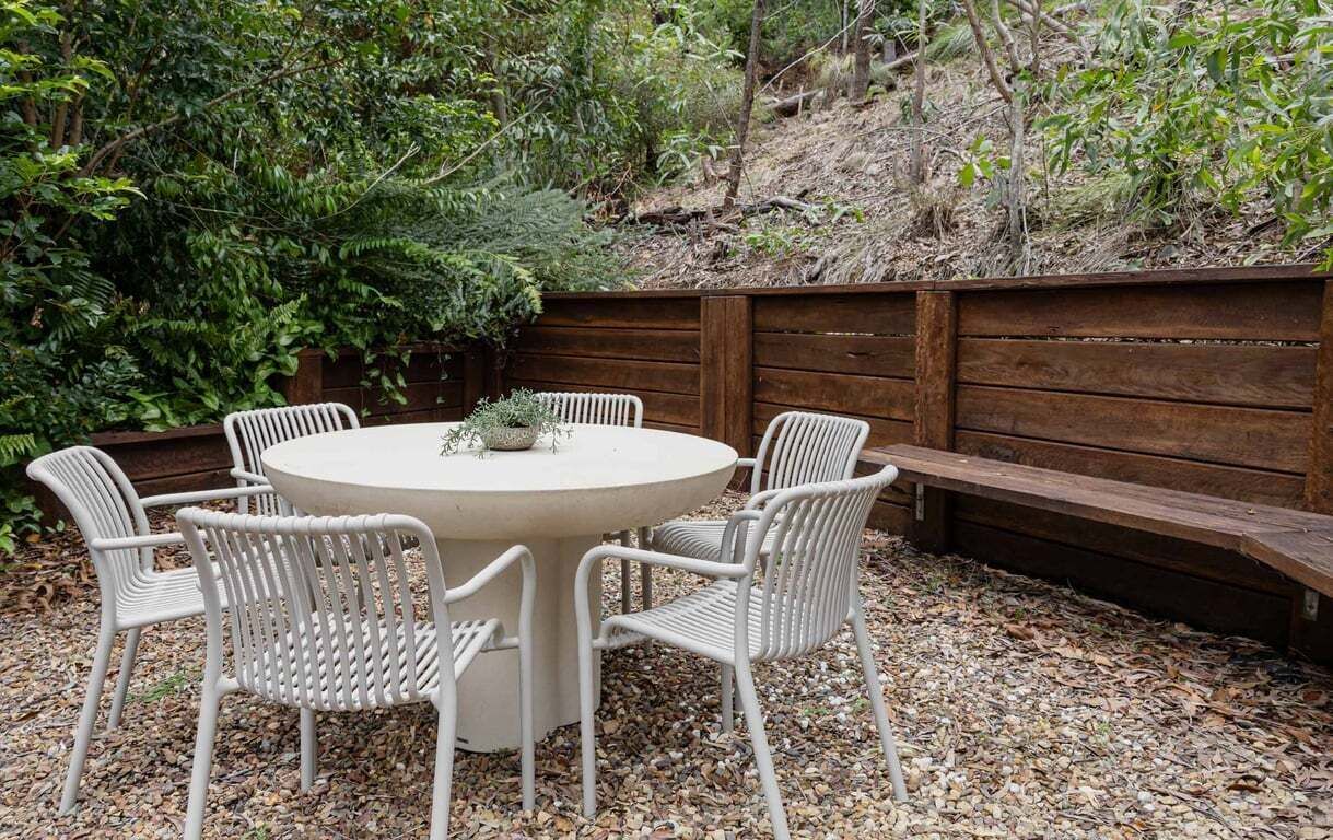A round outdoor table with six white chairs, surrounded by a wooden fence and dense foliage.