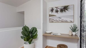 Een hoek van een kamer met een witte muur, een houten vloer, een potplant, een houten kruk, een plank met decoratieve objecten, en een lijst met een afbeelding van een strand.