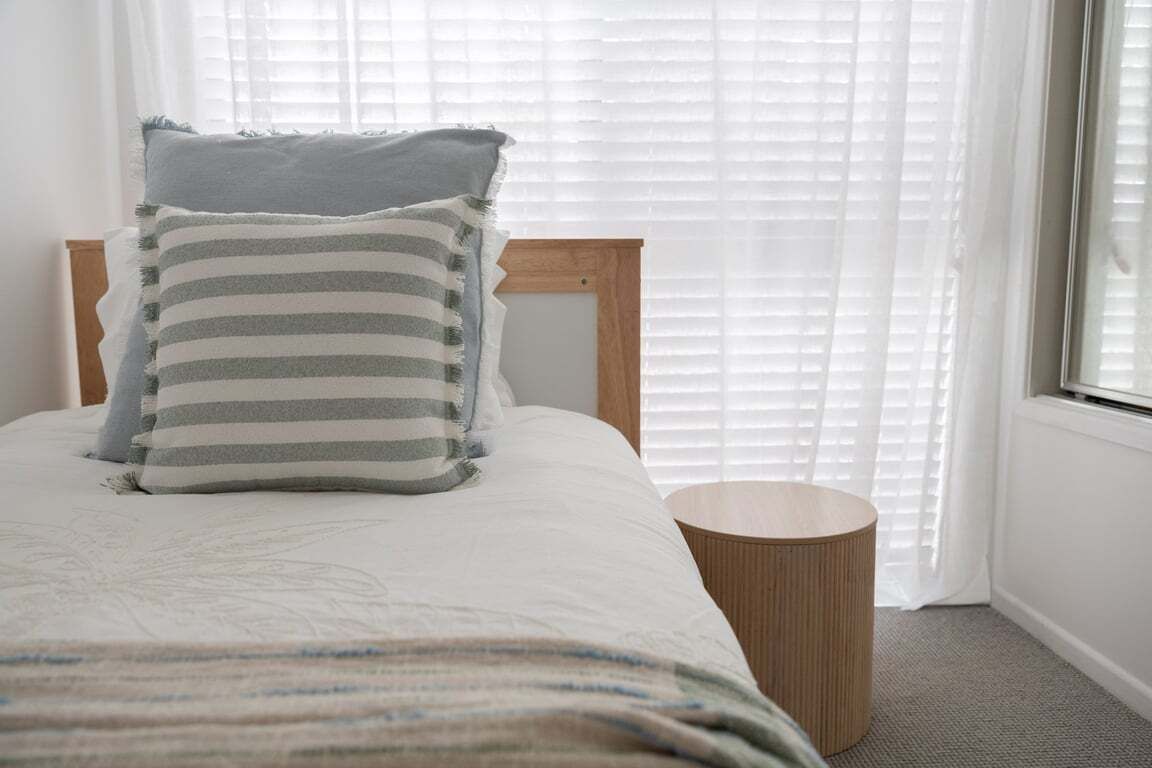 Een net opgemaakt bed met gestreept beddengoed en een houten hoofdbord, een rond houten nachtkastje ernaast, en een raam met jaloezieën op de achtergrond.