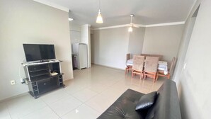 A living room with a black sofa, a TV on a stand, a dining table with chairs, and a ceiling fan. - Beautiful apartment located on Bombas Beach 🐶️ Sleeps up to 8 people (Bombinhas)