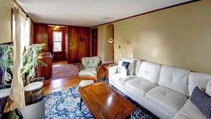 Ein Wohnzimmer mit einem weißen Sofa, einem gemusterten Teppich, einem Holztisch und einem karierten Sessel, sowie einem Topf mit einem Farn.