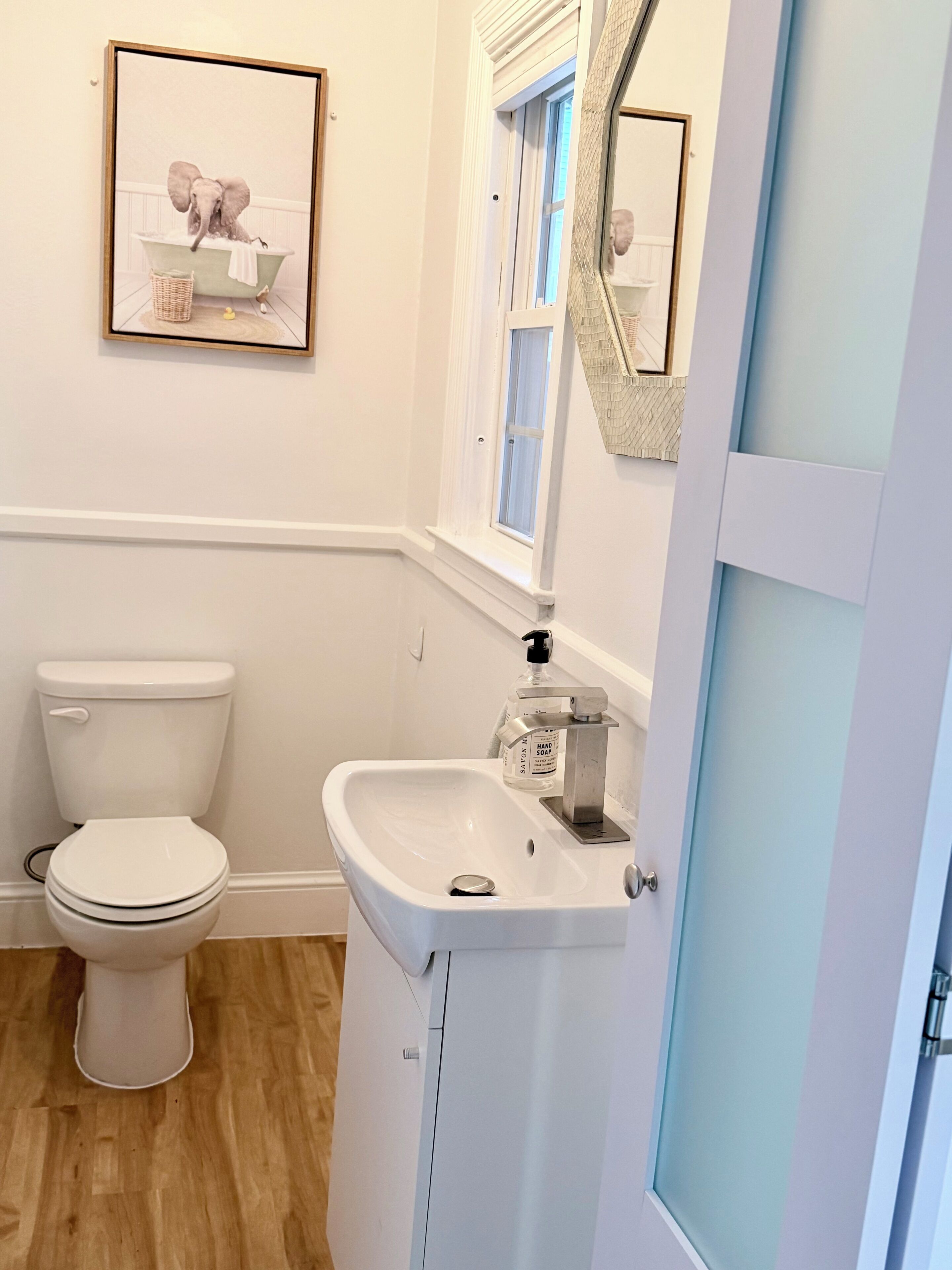 Une salle de bain avec des toilettes blanches, un lavabo blanc doté d’un distributeur de savon et un tableau encadré au mur.