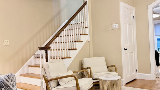 Una escalera con balaustres blancos y barandales de madera que conduce a un rellano con dos sillones tapizados y una pequeña mesita de madera.