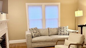Una sala de estar con un sofá beige, una mesa de centro de madera, un taburete blanco mullido y una lámpara.