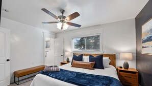 Une chambre à coucher comprenant un lit, un ventilateur de plafond, une fenêtre avec des stores, un banc et une table de chevet avec une lampe.