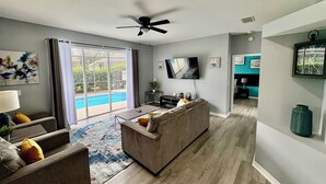 Ein modernes Wohnzimmer mit grauem Sofa, Couchtisch und Flachbildfernseher. Es gibt einen Deckenventilator, ein großes Fenster mit Vorhängen und einen Blick auf einen Swimmingpool.
