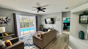 A modern living room with a grey sofa, a coffee table, and a flat-screen TV. There is a ceiling fan, a large window with curtains, and a view of a swimming pool.