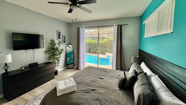 Ein Schlafzimmer mit Bett, Fernseher, Deckenventilator, Kommode und Blick auf einen Swimmingpool.