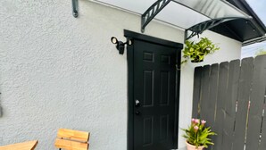 Un banc en bois, une porte noire, un paillasson avec l’inscription « BONJOUR » et une plante en pot.