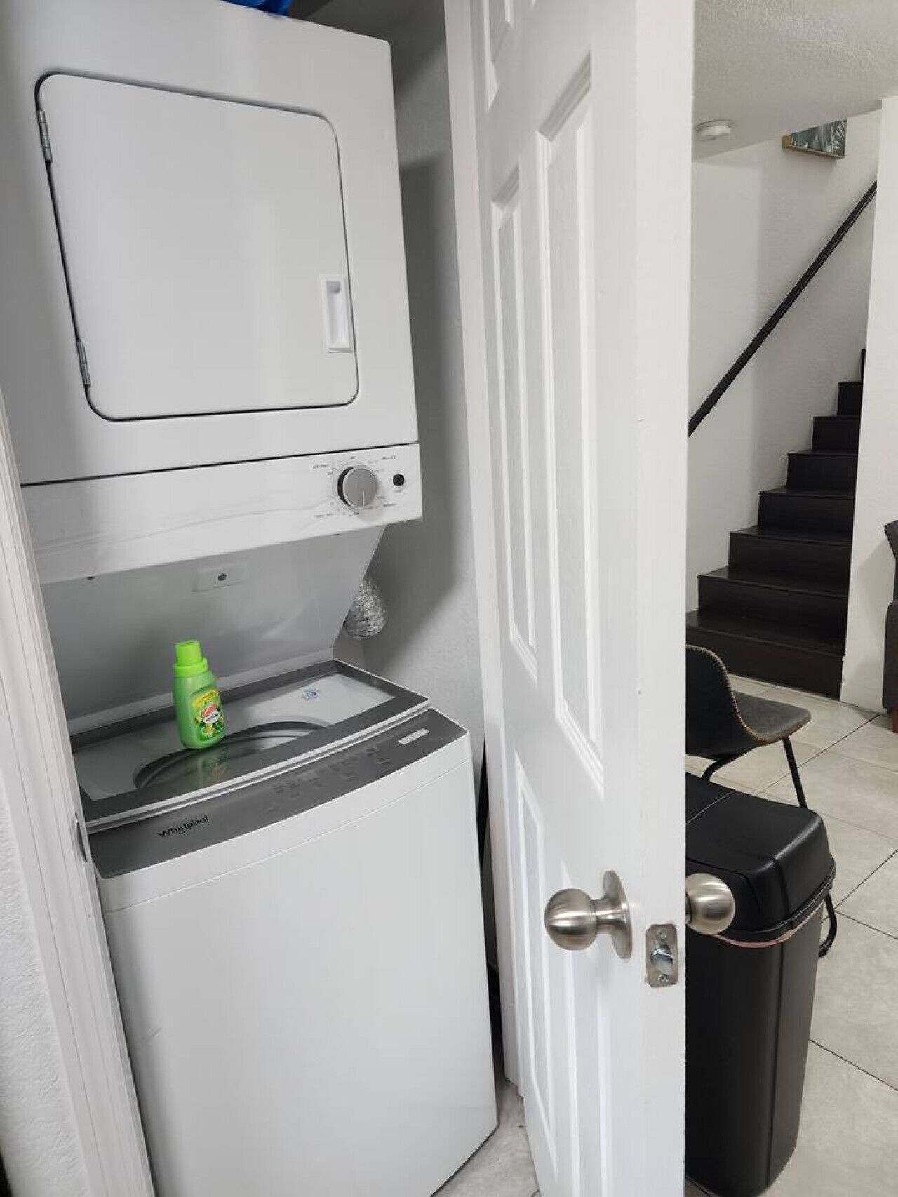Un área de lavandería compacta con lavadora y secadora, un estante pequeño con una botella, y una escalera al fondo.