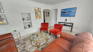 Un salon avec un canapé en cuir marron, une table basse en verre, deux fauteuils rouges et un téléviseur fixé au mur.