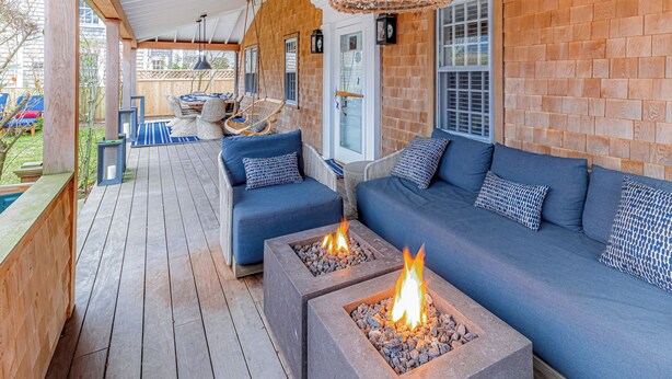 Ein überdachter Außenbereich mit Holzdeck, blauen Sofas und zwei quadratischen Feuerstellen mit Flammen.