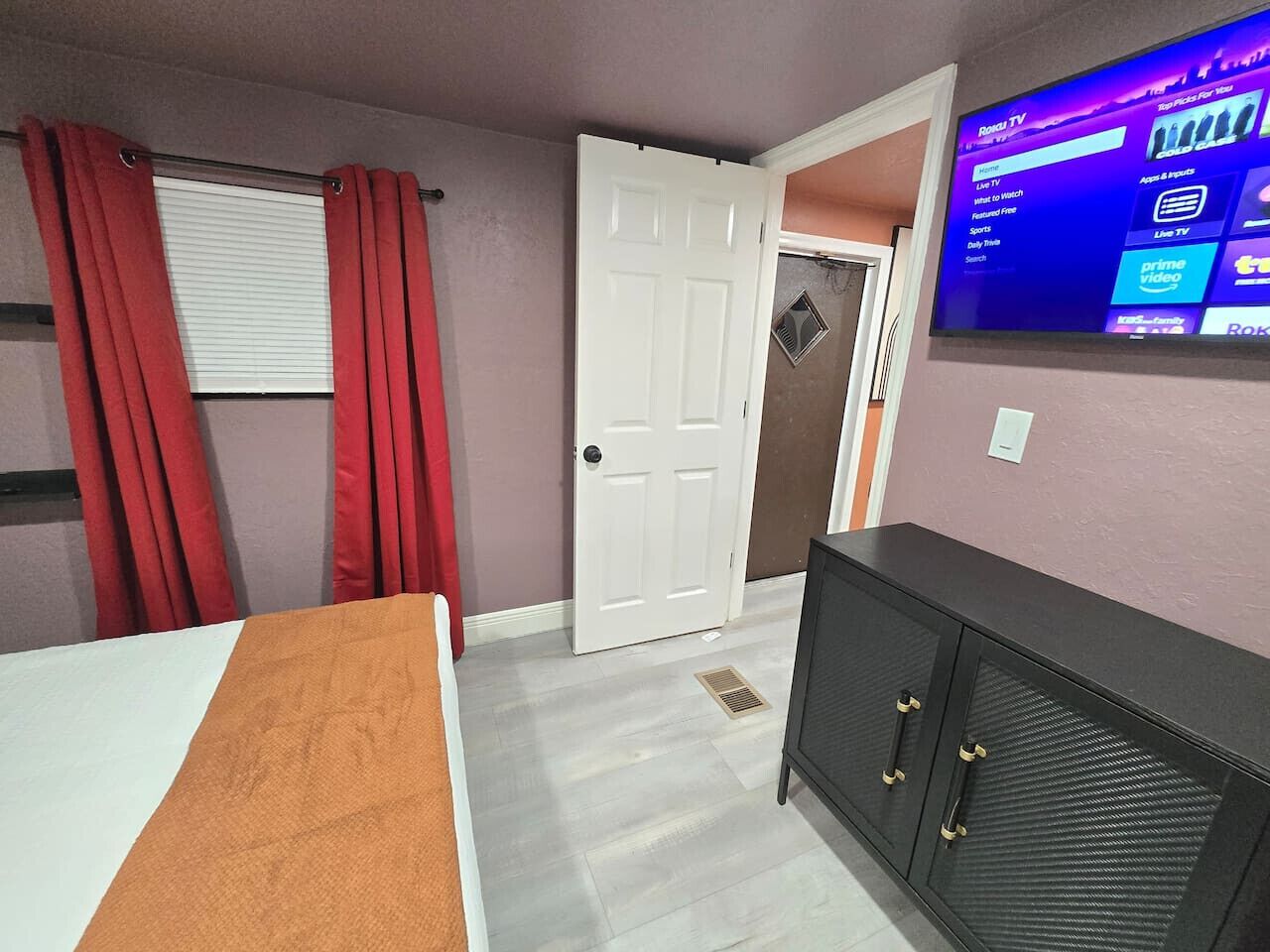 A room with a bed, a TV mounted on the wall, a door, and a window with red curtains.