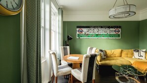 Una stanza con una parete verde, un divano giallo, sedie a righe, un tavolo rotondo in legno e un lampadario.