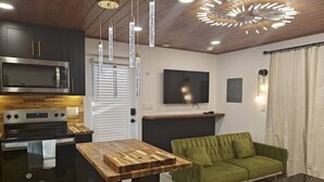 Cozinha moderna com micro-ondas, fogão e mesa de madeira. Há um sofá verde e uma TV de tela plana na parede.