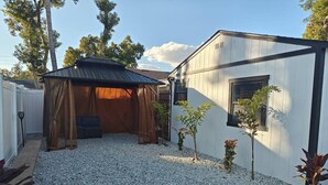 Uma área externa coberta com chão de cascalho, um pequeno galpão e um edifício branco com uma janela.