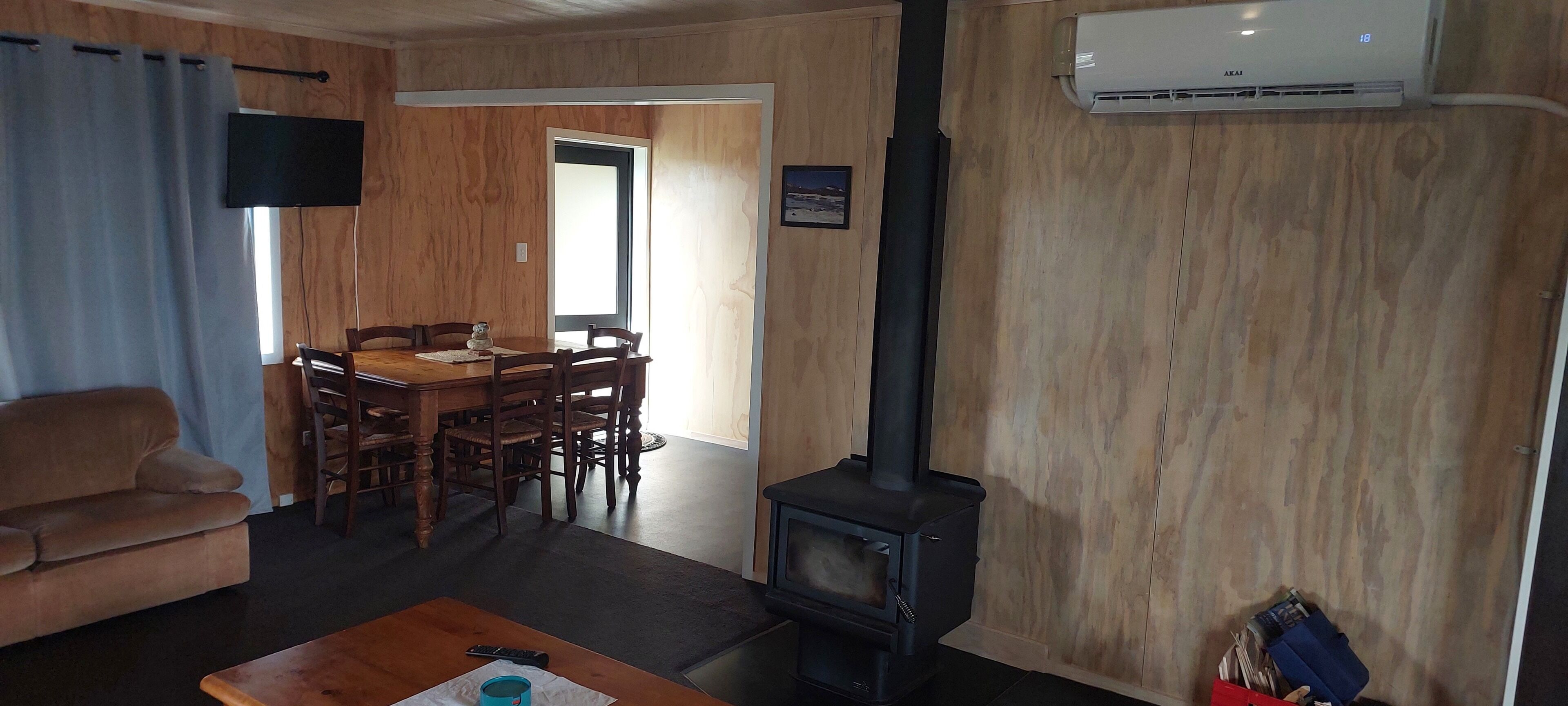 A living room with a sofa, a wooden dining table with chairs, a black wood-burning stove, and a mounted TV.