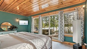 A bedroom with a wooden ceiling, a bed with a patterned bedspread, a large window with curtains, and a view of the outdoors. - Spacious 5-Bedroom -Hot Tub, MDI, Lake front, Fire pit, Game Room, Dog Friendly! (Franklin)