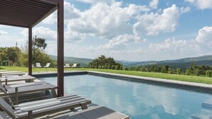 Una piscina con lettini e un pergolato, immersa nel verde e circondata da colline.