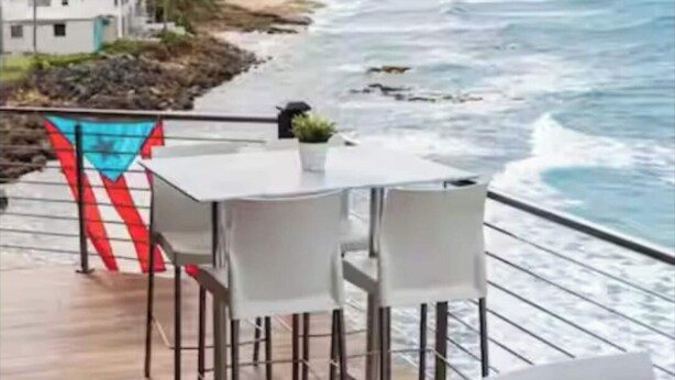 Una terraza con sillas blancas y una mesa con vista al océano, donde se ve una playa y un edificio a lo lejos.