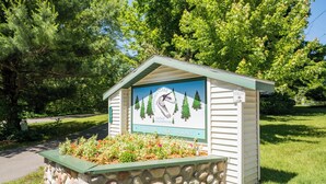 A small stone-built cottage with a signboard depicting a mountain and trees, surrounded by a flower bed and greenery. - Goshorn Lake #1! Beach Access - Shared Pool - 1 min from Dune Rides (Saugatuck)