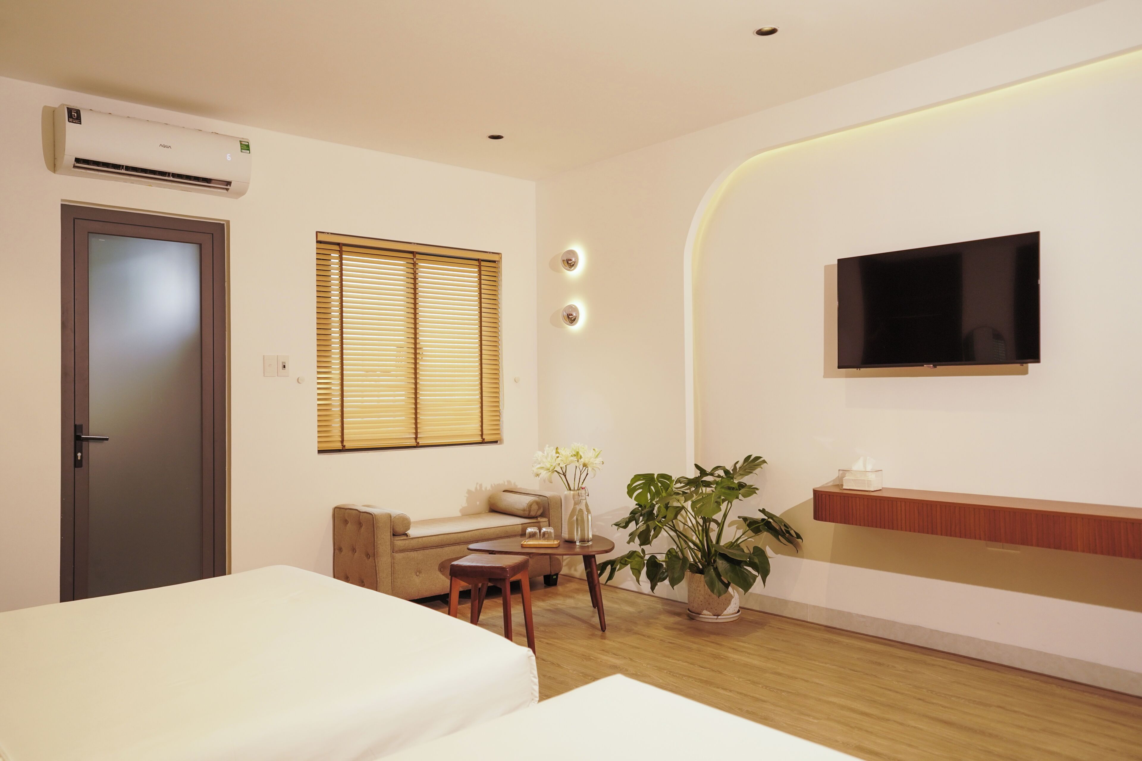 A hotel room with a bed, a sofa, a small table with a vase of flowers, a potted plant, a TV mounted on the wall, and a wooden floor.