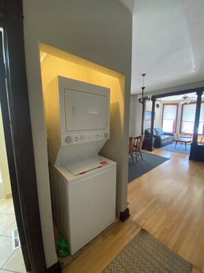Een wasserette met een wasmachine en droger, een houten vloer, en uitzicht op een woonruimte met een bank en een tafel.