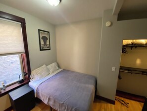 A bedroom with a bed, a desk with a lamp, a window with blinds, a wall-mounted shelf, and a door. - Sweet 2-bedroom + den apartment with AC in vibrant Bucktown (Chicago)