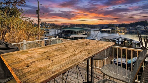 A wooden table and metal chairs on an outdoor deck with a view of a river and buildings in the distance. - Charming Lakefront House Near Shorty Pants at 21mm (Osage Beach)