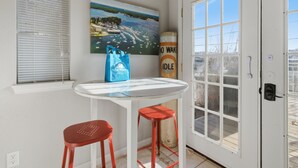 A small room with a white door, a window with blinds, a painting of a coastal scene, a blue bag, and two red stools. - Charming Lakefront House Near Shorty Pants at 21mm (Osage Beach)