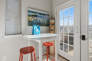 A small room with a white door, a window with blinds, a painting of a coastal scene, a blue bag, and two red stools. - Charming Lakefront House Near Shorty Pants at 21mm (Osage Beach)