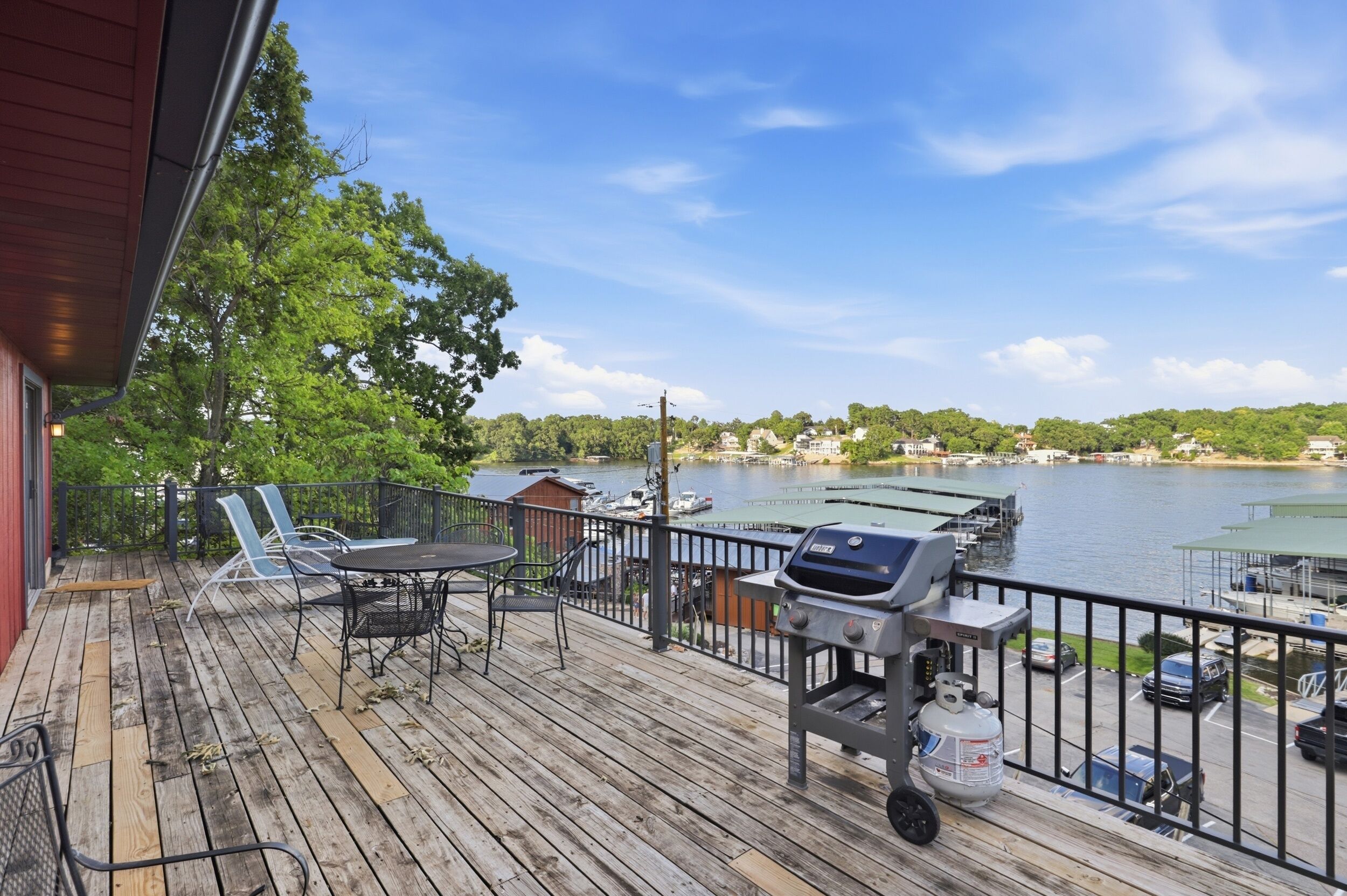 Une terrasse en bois, meublée pour l’extérieur, avec un barbecue et une vue sur un lac, où l’on aperçoit des bateaux et des bâtiments au loin.