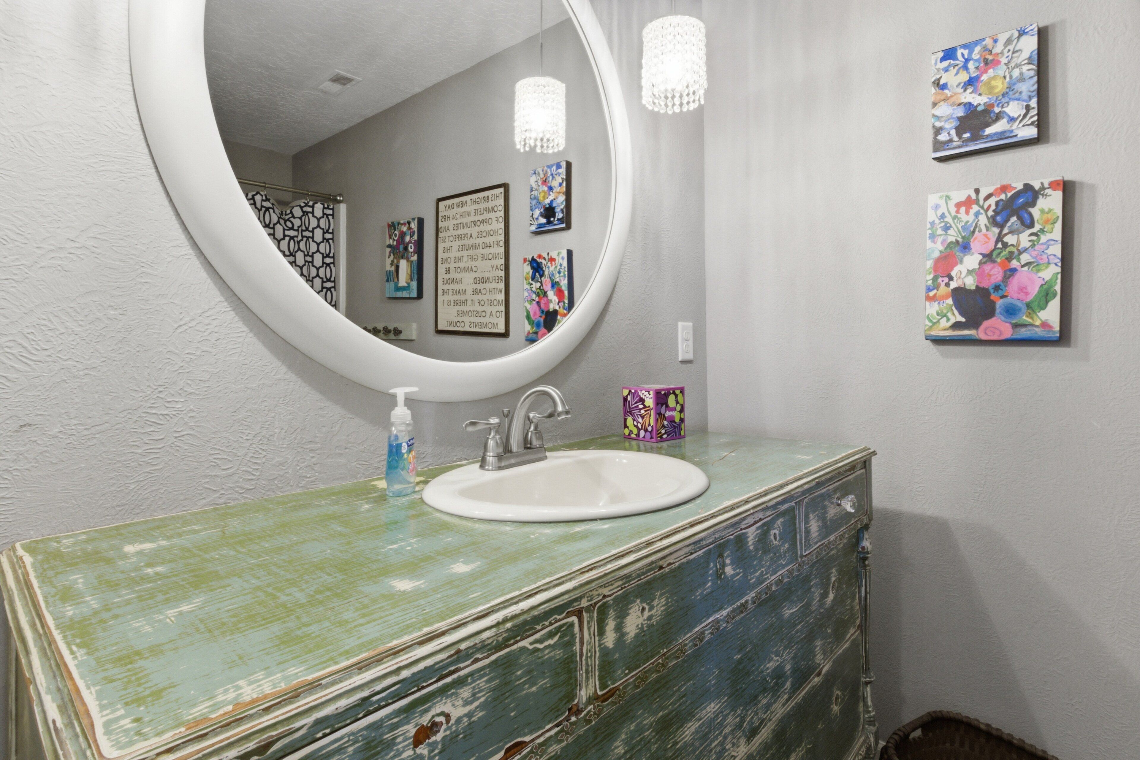 Un baño con un espejo redondo, un lavabo blanco y un aparador azul verdoso.