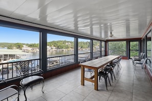 A covered outdoor area with a long table and chairs, large windows with a view of a body of water and buildings, and a tiled floor. - Lakefront Home w/ Decks & Docks at Shorty Pants Bar 21mm (Osage Beach)