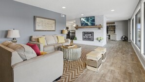Ein geräumiges Wohnzimmer mit Sofa, Couchtisch und Pouf. Ein Kamin mit einem dekorativen Schild darüber. Im Hintergrund ist ein großer Fenster und ein Blick in die Küche zu sehen.