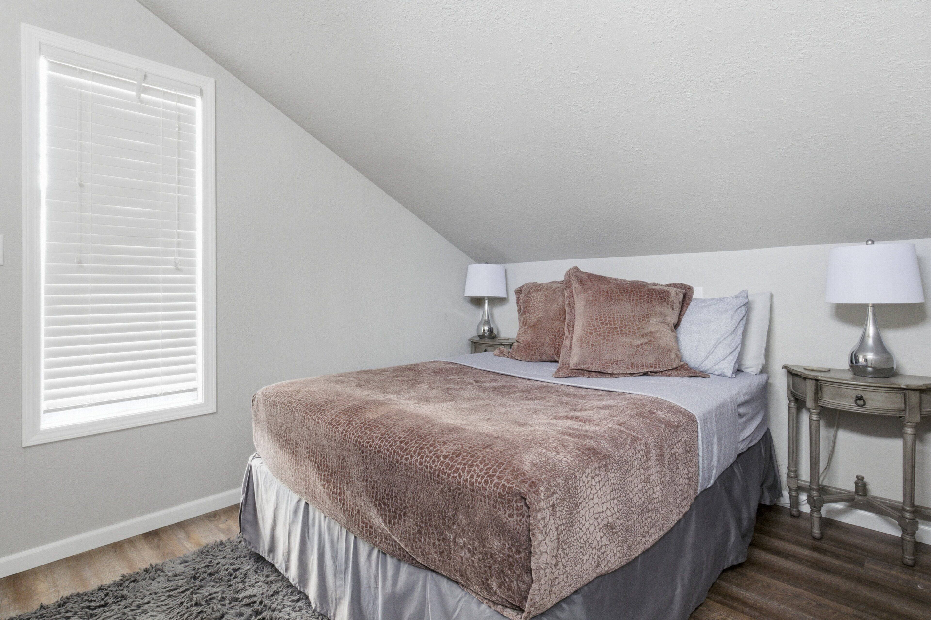 A bedroom with a bed, two bedside lamps, a window with blinds, and a nightstand.