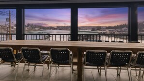 Ein Konferenzraum mit großem Tisch und Stühlen, großen Fenstern mit Blick auf einen Fluss und Gebäude sowie einer Metallreling.