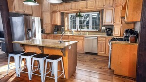 Une cuisine avec des meubles de rangement en bois, un réfrigérateur en acier inoxydable, un évier, une cuisinière et trois tabourets de bar.