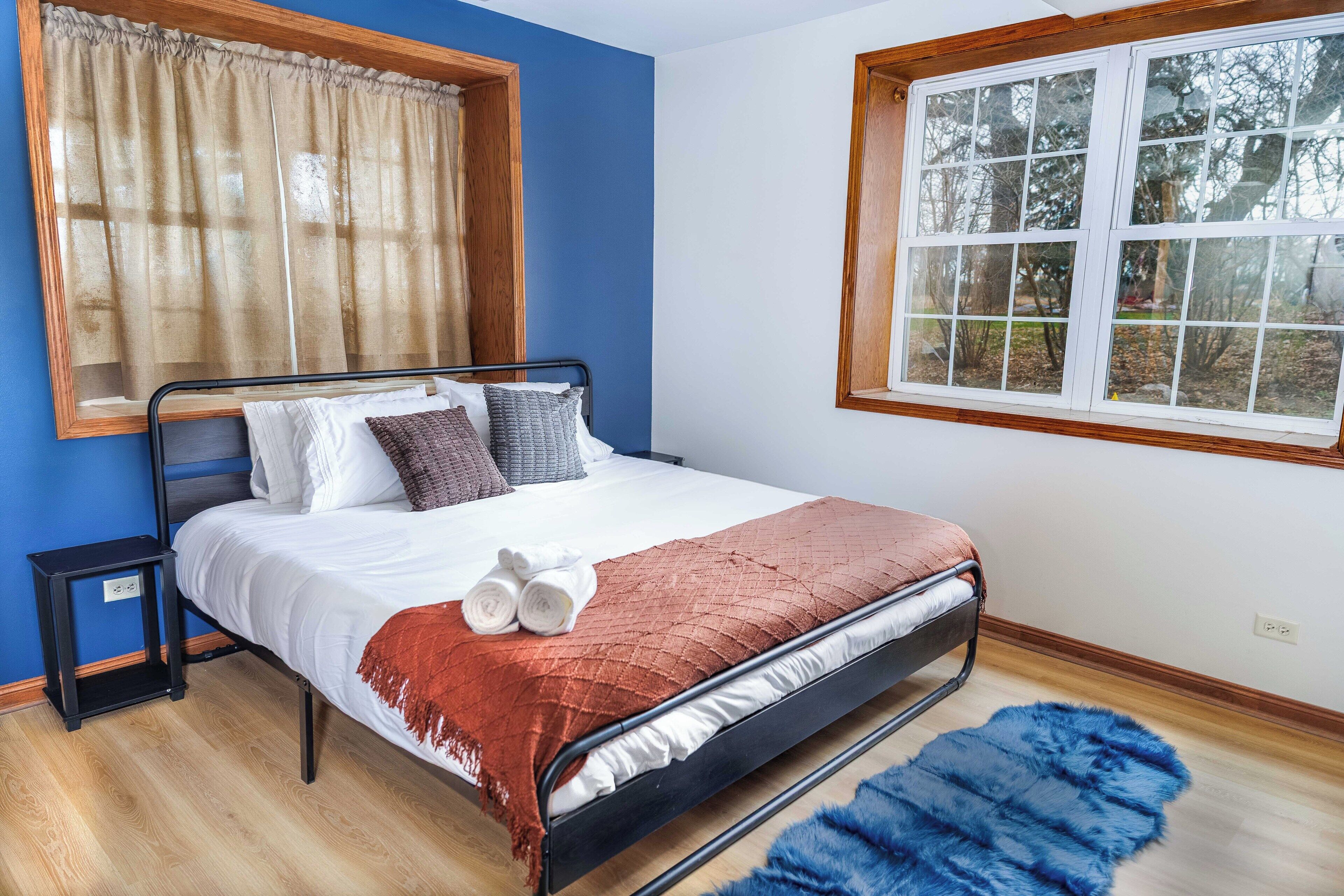 A bedroom with a bed, a nightstand, a window with a view of trees, and a blue rug.