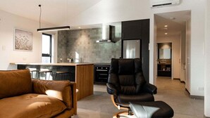 Een moderne woonkamer met een leren bank, een zwarte leren fauteuil en een houten salontafel. Achtergrond toont een keuken met donkere kastjes en een roestvrijstalen koelkast.