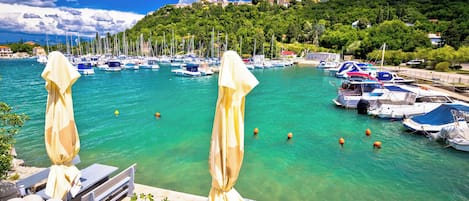 Ein Yachthafen mit Booten, ein Holzdeck mit Stühlen und Sonnenschirmen sowie eine Hügelside mit Gebäuden im Hintergrund.