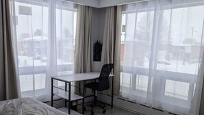 A bedroom with a bed, a desk, and two large windows with sheer curtains.