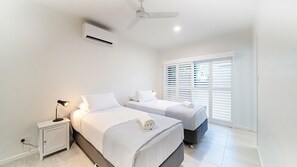 A bedroom with two beds, a ceiling fan, an air conditioning unit, a bedside table with a lamp, and a window with blinds. - Golden Tides - Mission Beach (Mission Beach)